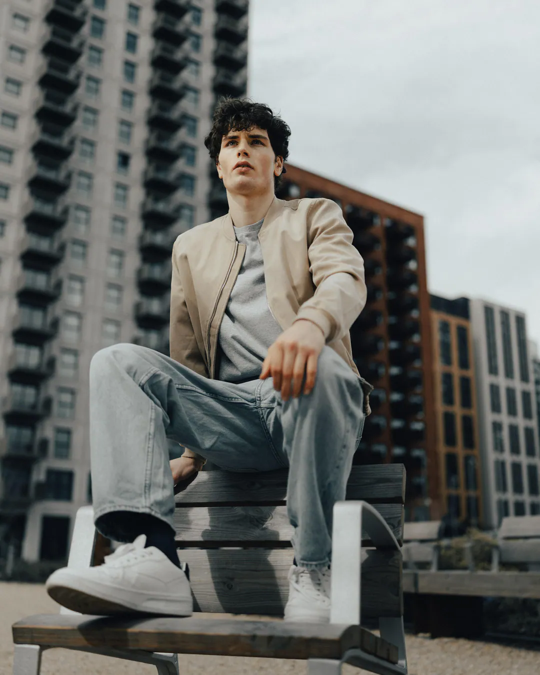 Man in a beige bomber jacket, pale jeans and white trainers perched on a bench below apartment blocks