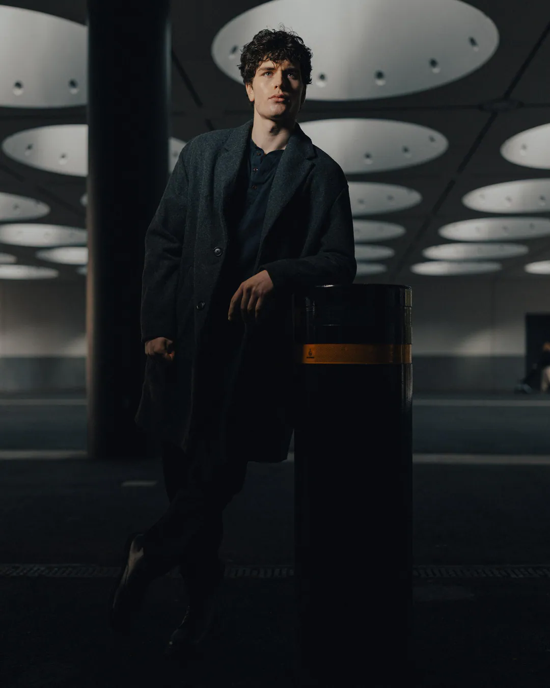 Man in a dark coat leaning against a black bollard beneath circular skylights in a shaded walkway