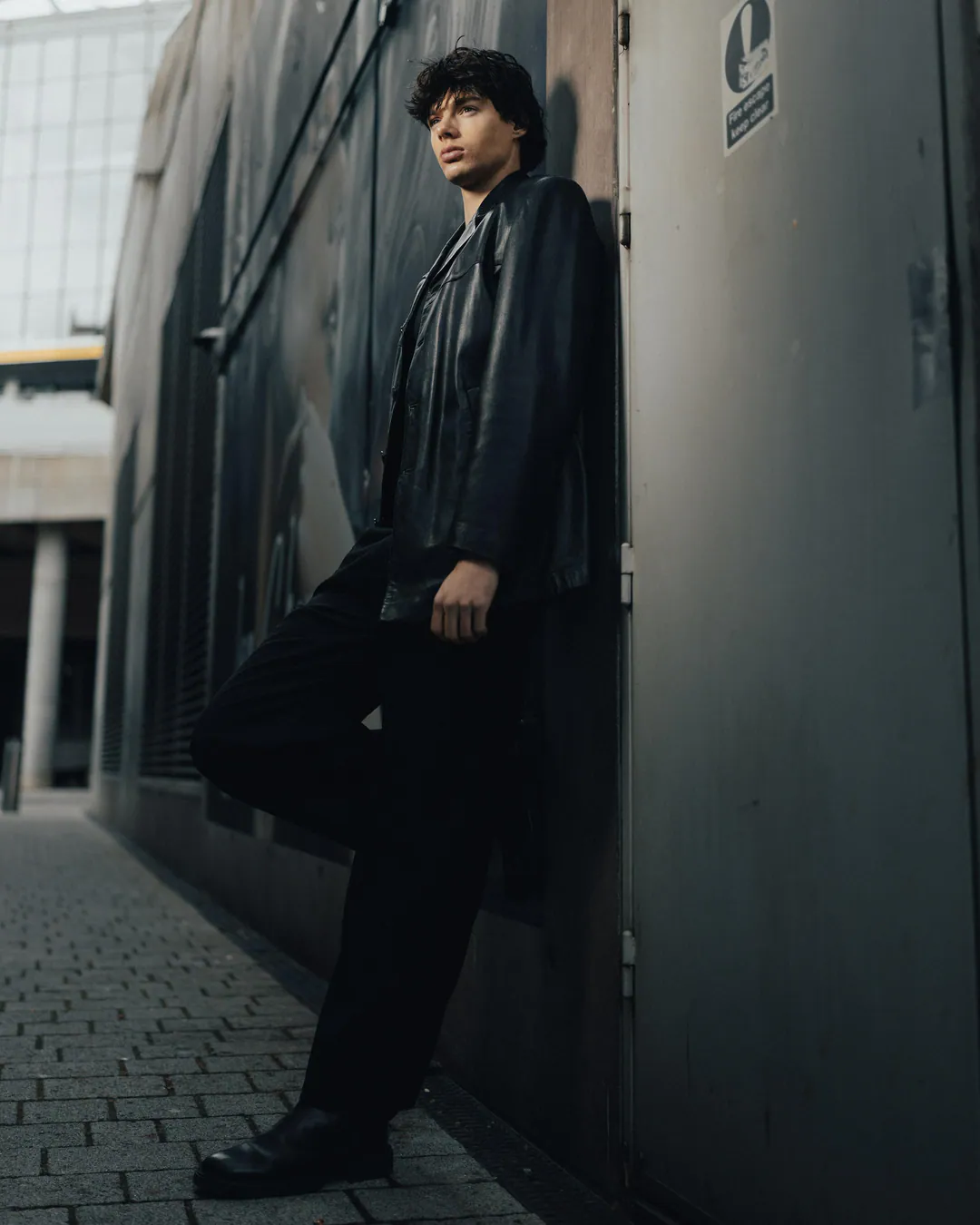Man in a black leather jacket leaning against a metal wall in a narrow paved alleyway