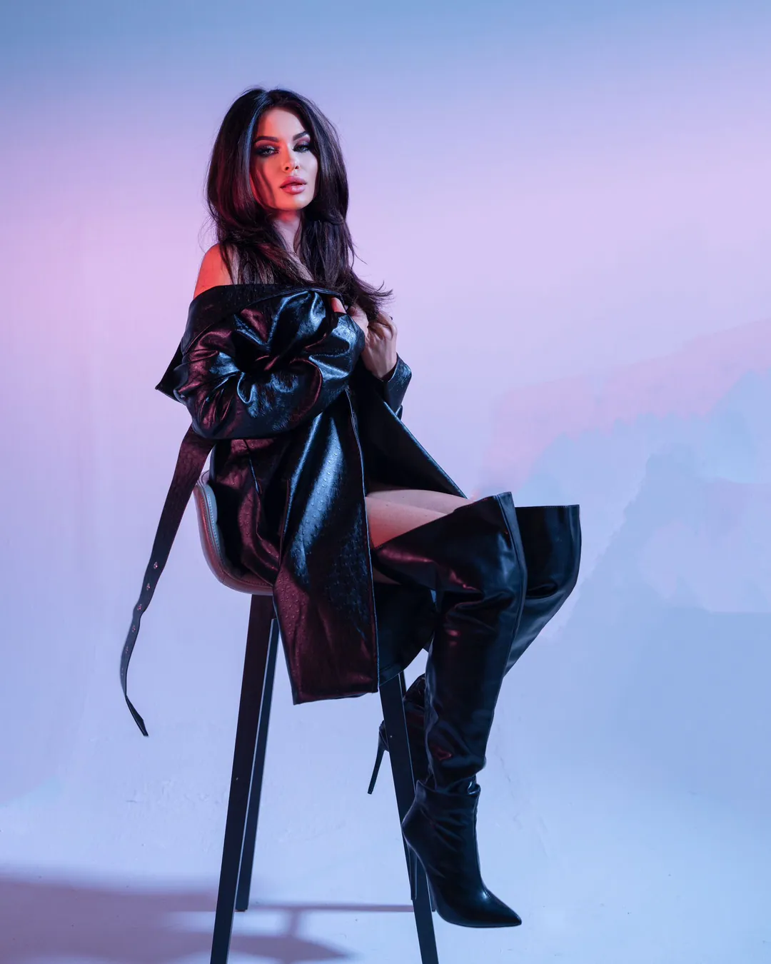 Woman in a leather coat and thigh-high boots seated on a stool against a pastel lilac backdrop
