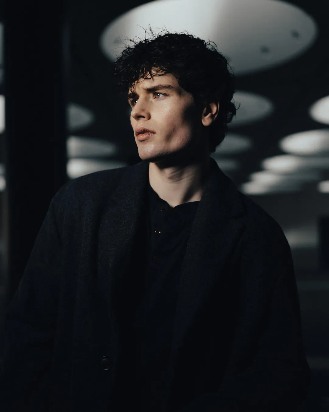 Man in a dark wool coat glancing sideways in low light beneath a ceiling of circular skylights
