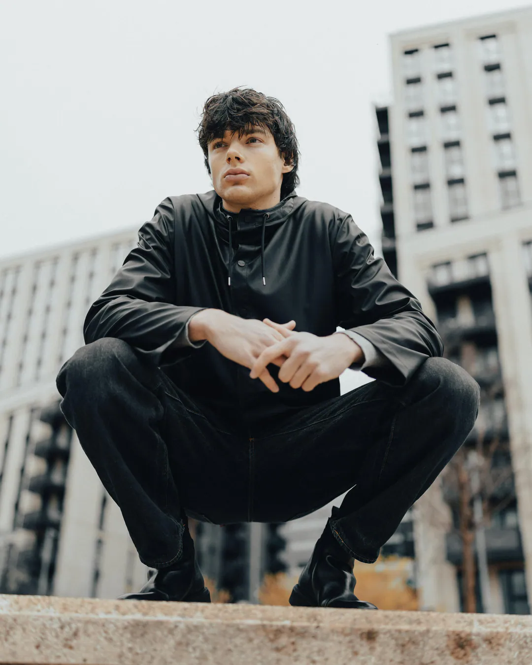 Man in a black leather jacket and trousers crouching on a stone ledge against modern apartment blocks