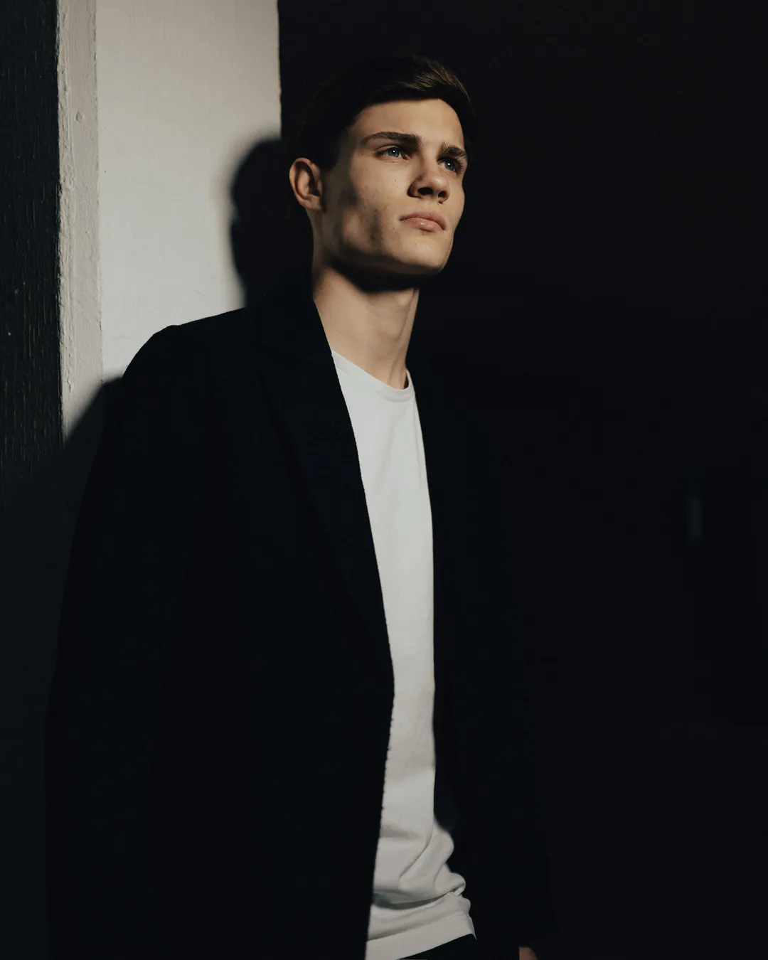 Man in a black coat and white T-shirt leaning against a pale pillar, his face lit against darkness