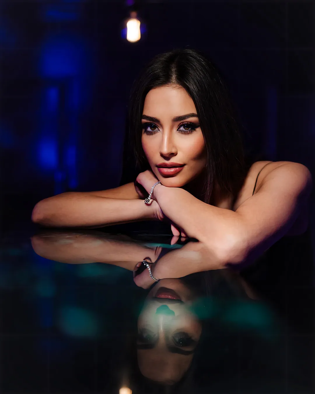 Woman with dark hair resting her chin on folded arms over a reflective table in deep blue light