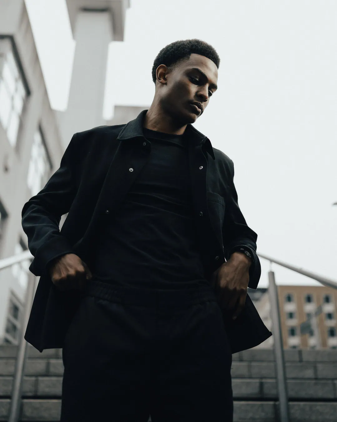 Man in a black overshirt and trousers standing on outdoor steps, looking down, against a pale sky