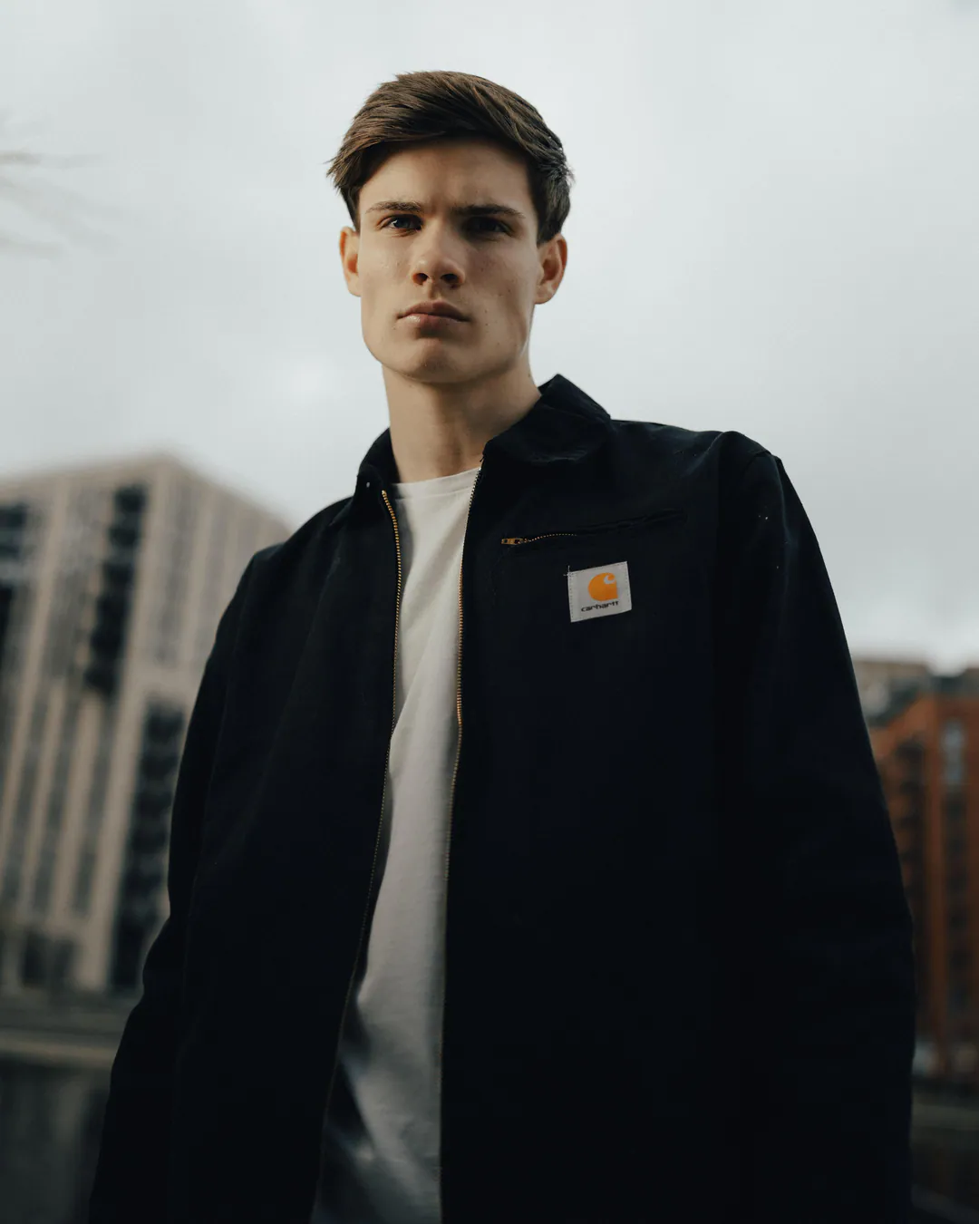 Man in a black workwear jacket and white T-shirt, shot from below against modern flats and grey sky
