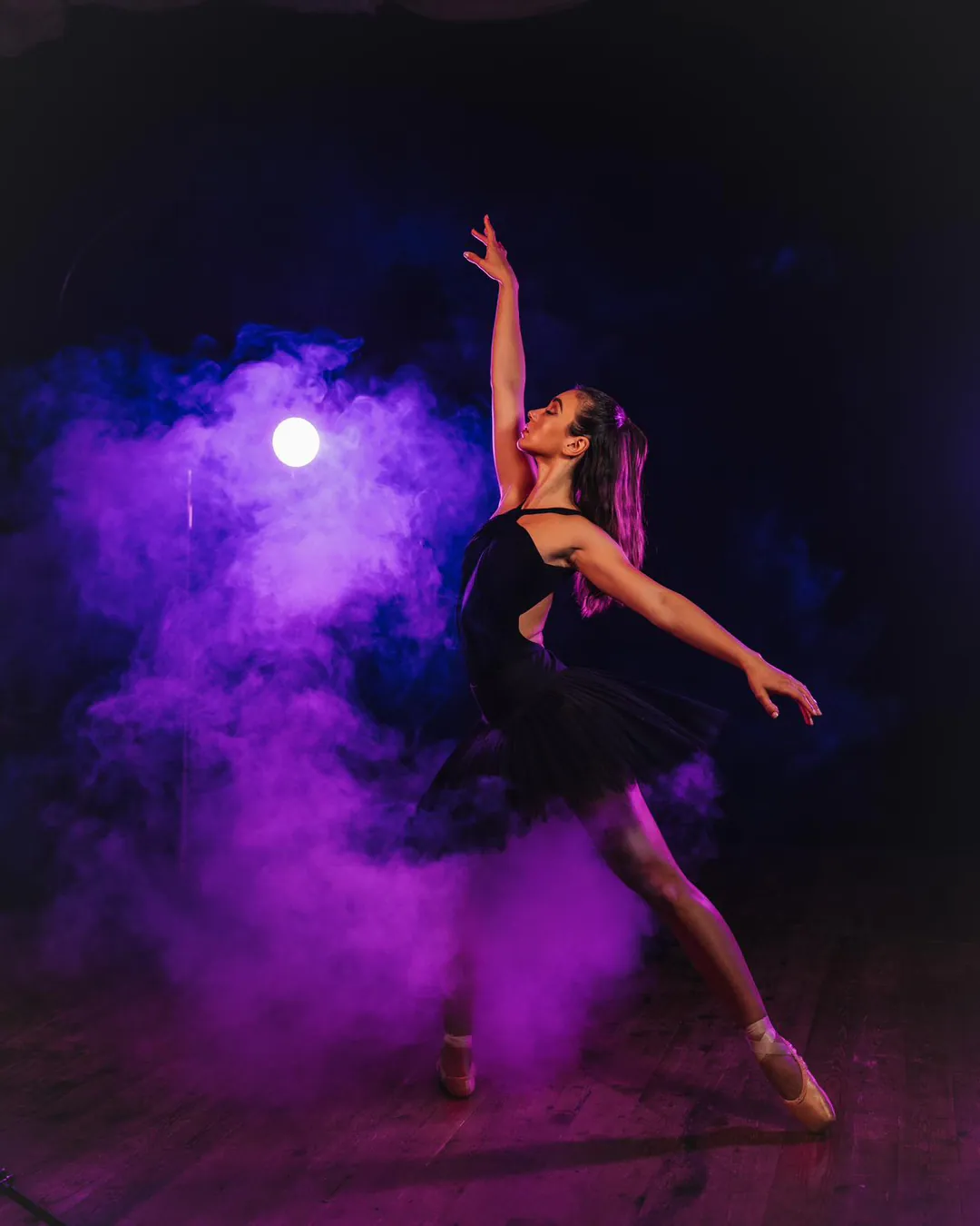 Ballet dancer in a black leotard and tutu posing en pointe amid purple smoke against a dark backdrop