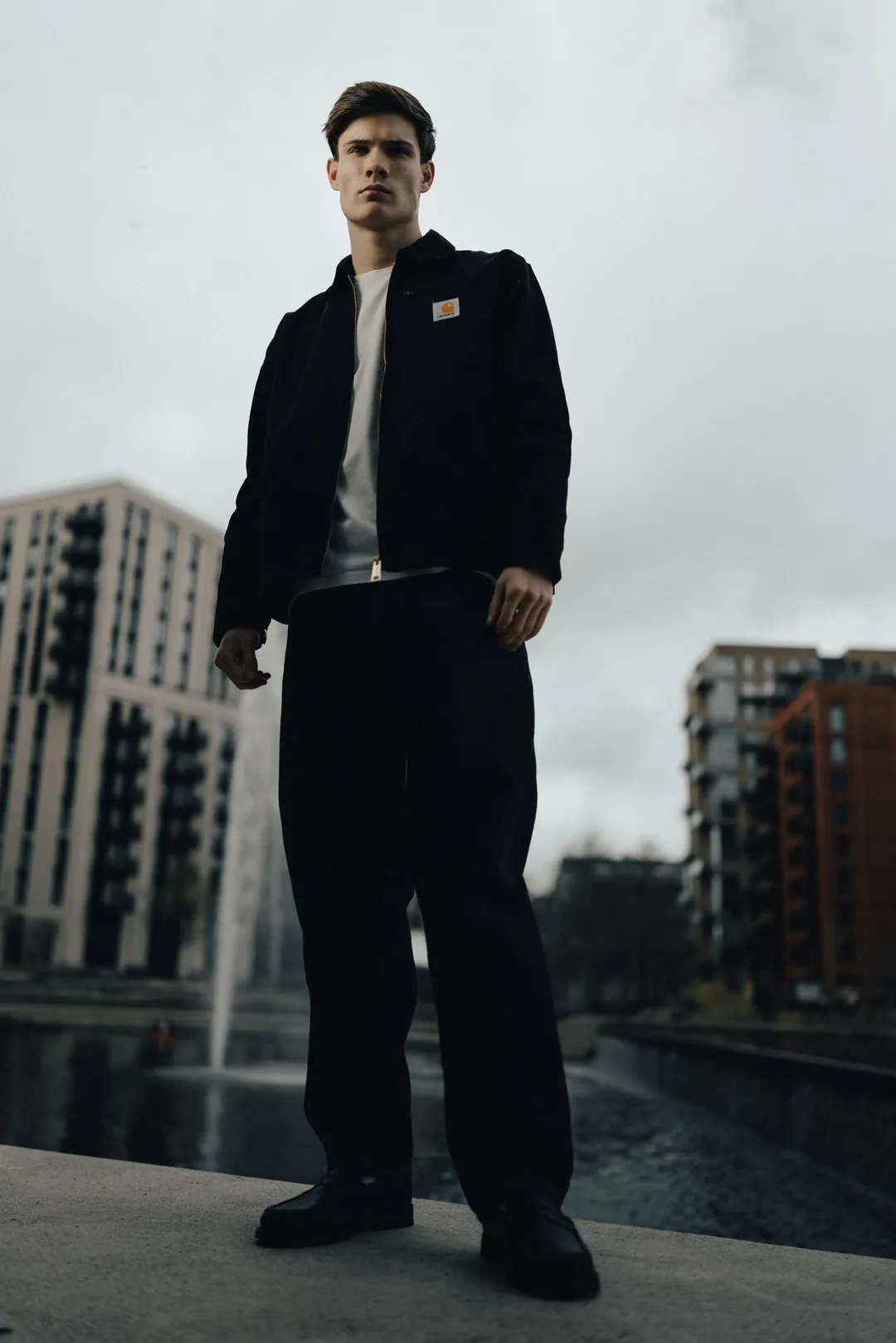 Man in a black jacket and wide black trousers standing on a stone ledge beside a fountain