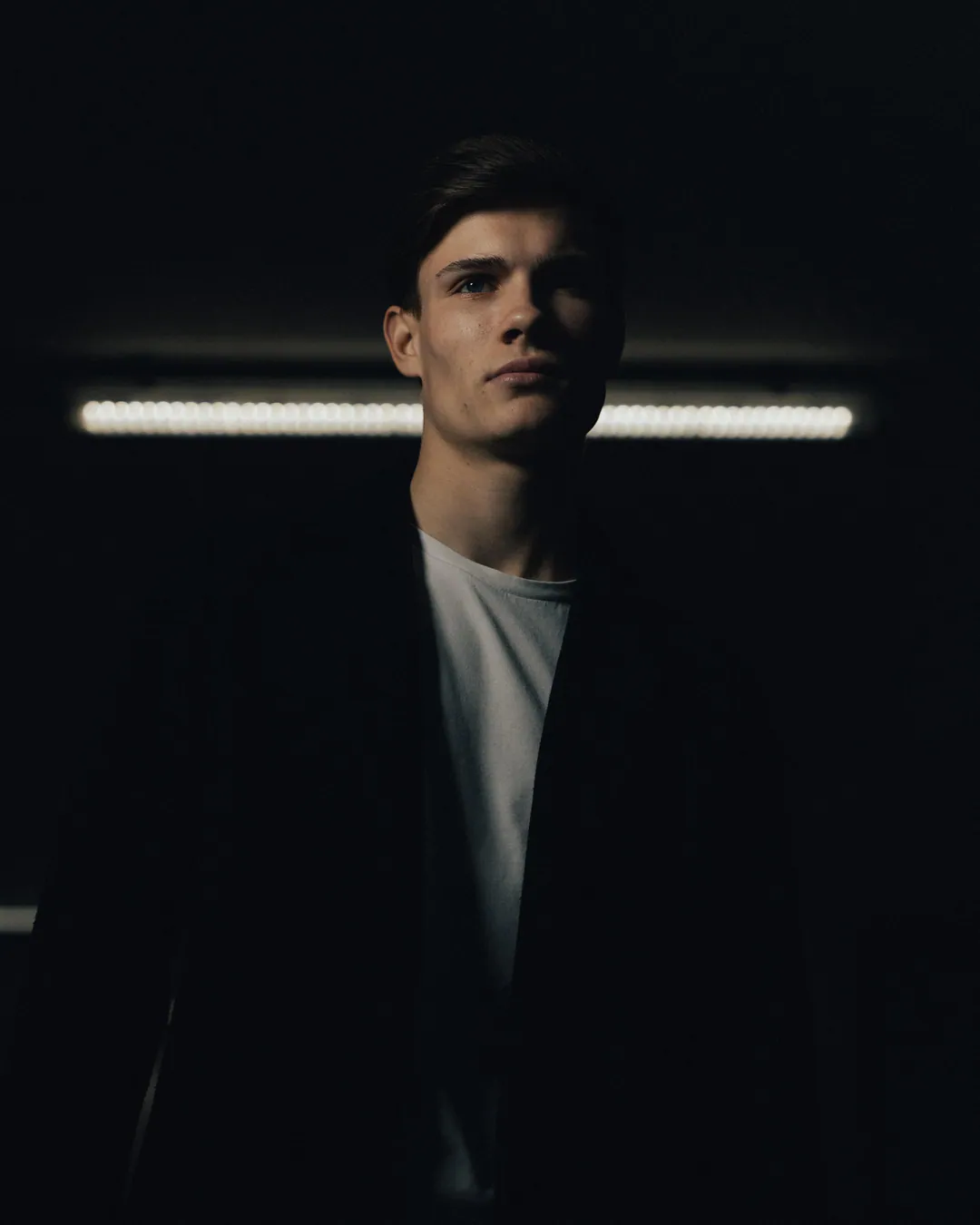 Man in a dark coat and white T-shirt in a dim underground space lit by a strip light