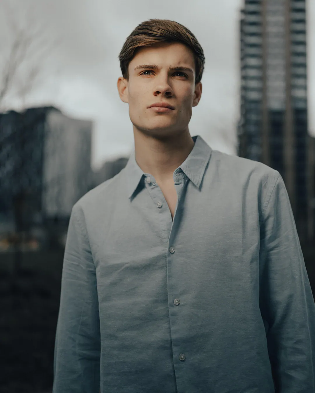 Man in a pale blue button-up shirt standing among high-rise buildings in soft light under a grey sky