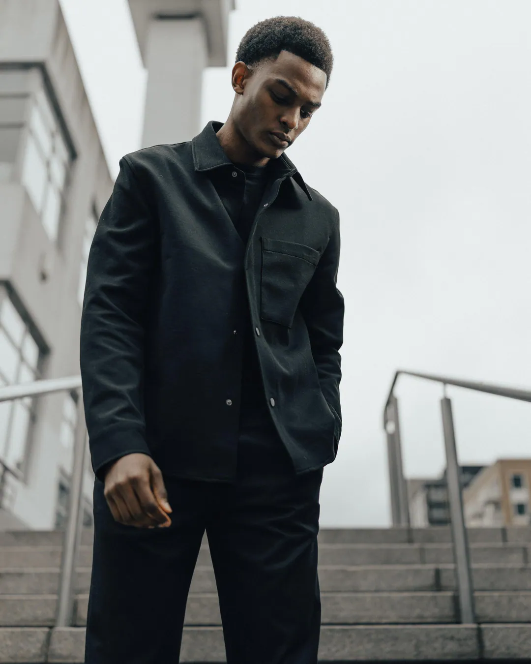 Man in a black overshirt standing on concrete steps with metal railings, gazing downwards, low-angle view