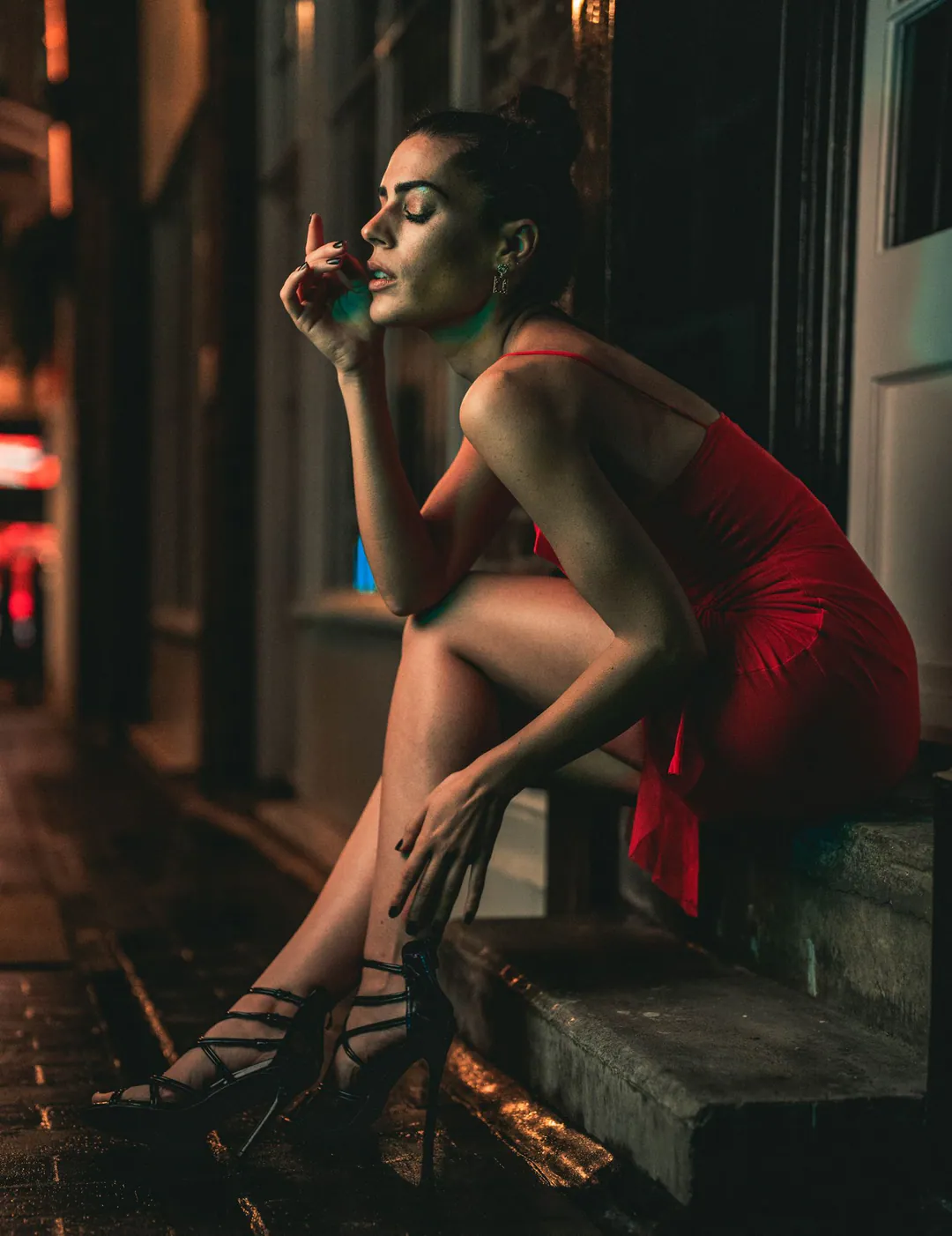 Woman in a red dress and strappy heels sitting on a step at night, eyes closed, hand raised