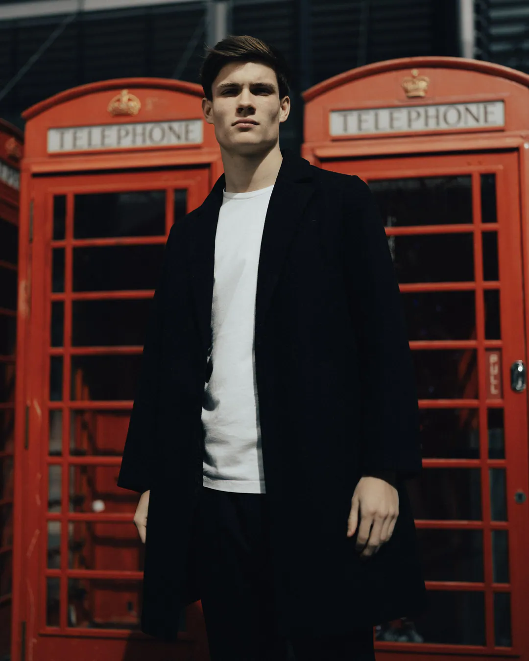Man in a long black overcoat and white T-shirt standing tall between two red telephone boxes