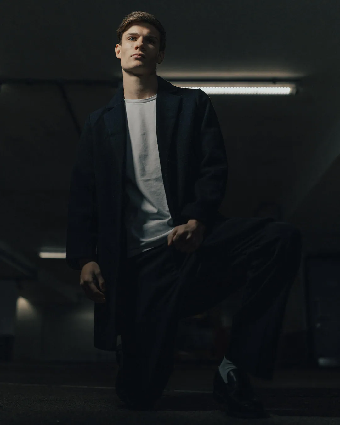 Man in a long black coat kneeling on one knee in a dark car park beneath strip lights