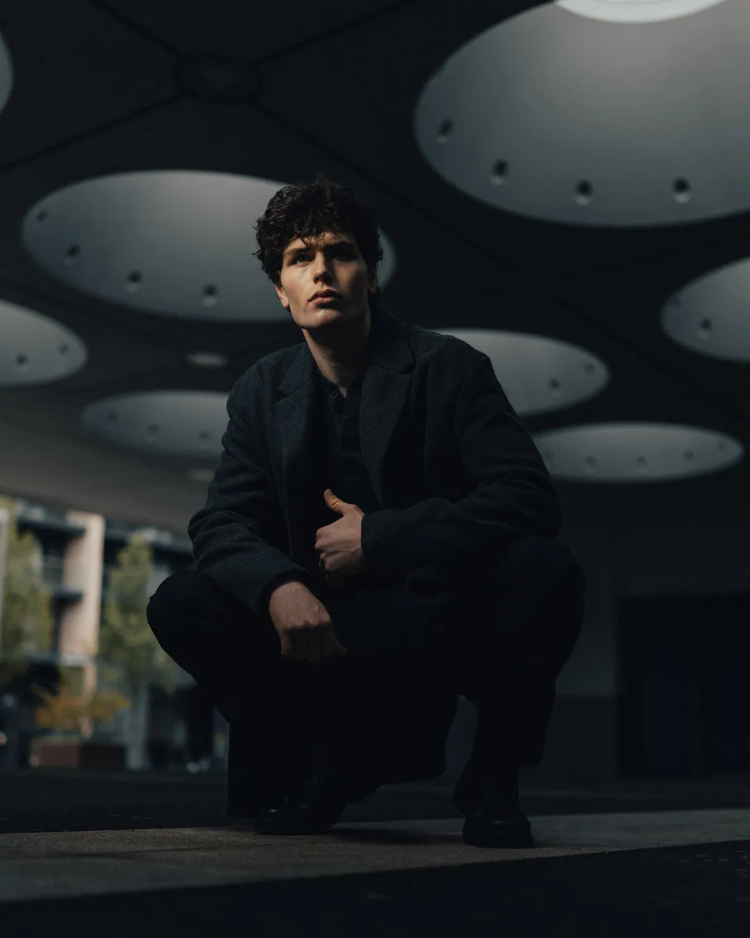 Man in a black jacket crouching low beneath a ceiling of circular skylights, looking past the camera