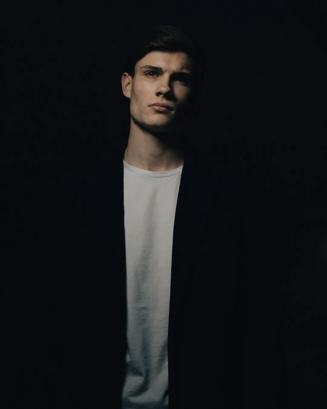 Man in a black coat and white T-shirt emerging from deep shadow, face caught in light