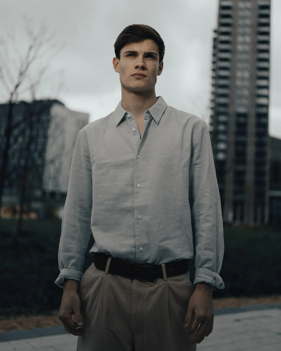 Man in a pale blue linen shirt and beige trousers standing on a city plaza beneath grey skies