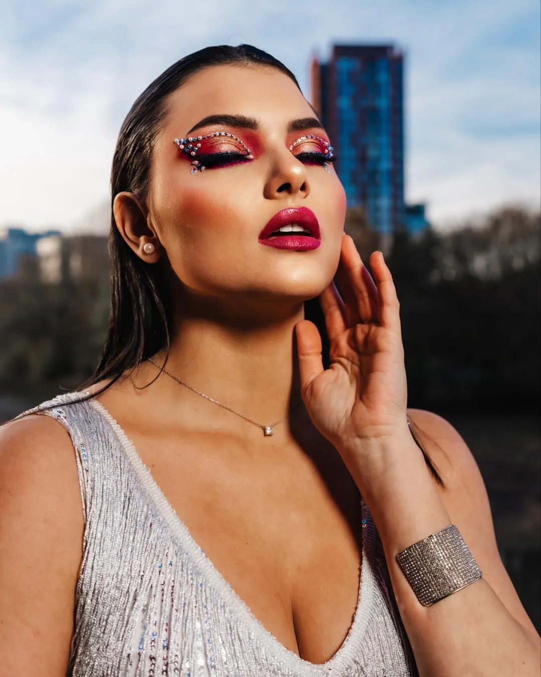 Woman in a silver sequinned top with glittering pink eye make-up, hand at her face, city towers behind
