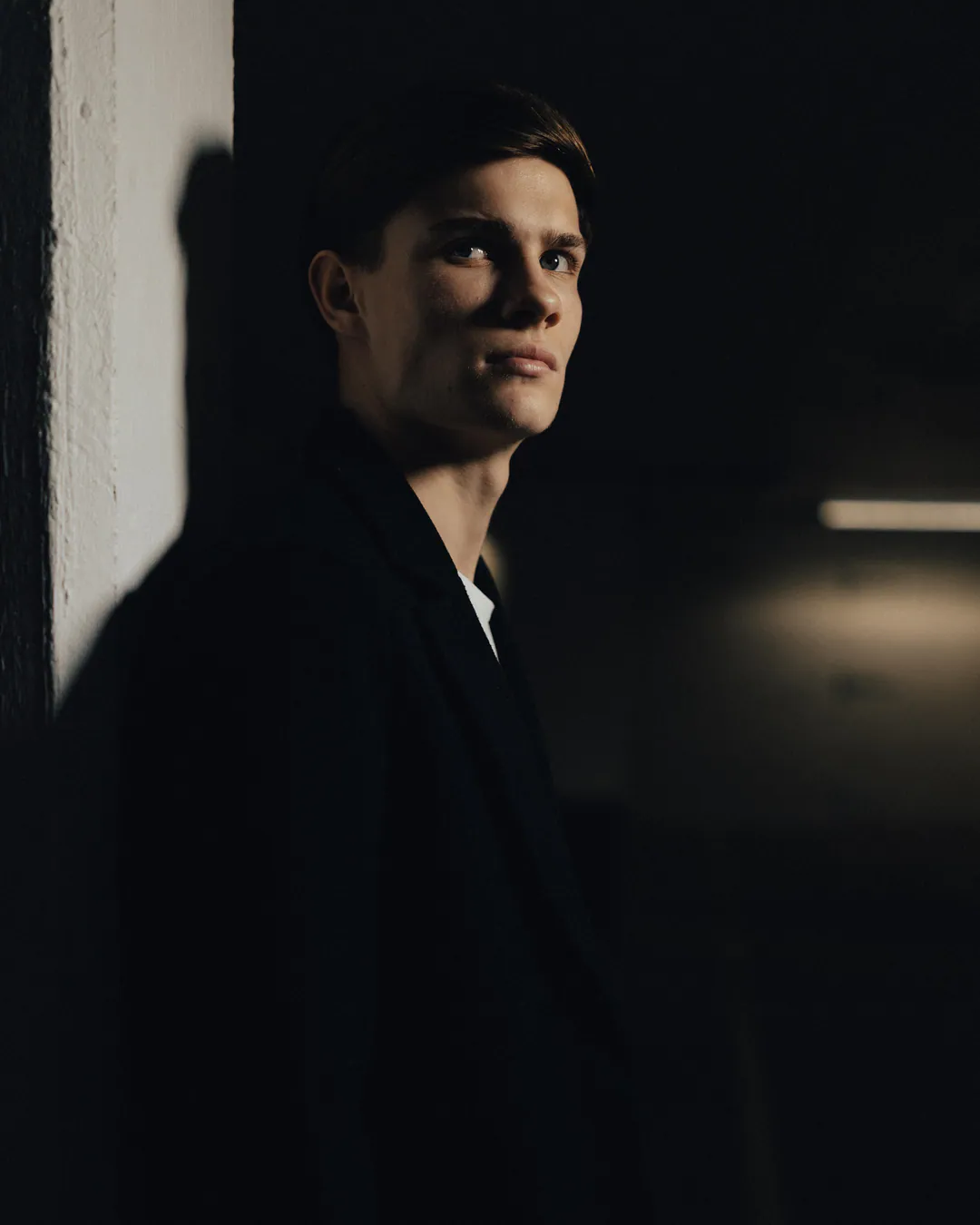 Man in a dark coat leaning against a white pillar in deep shadow, looking upwards to one side