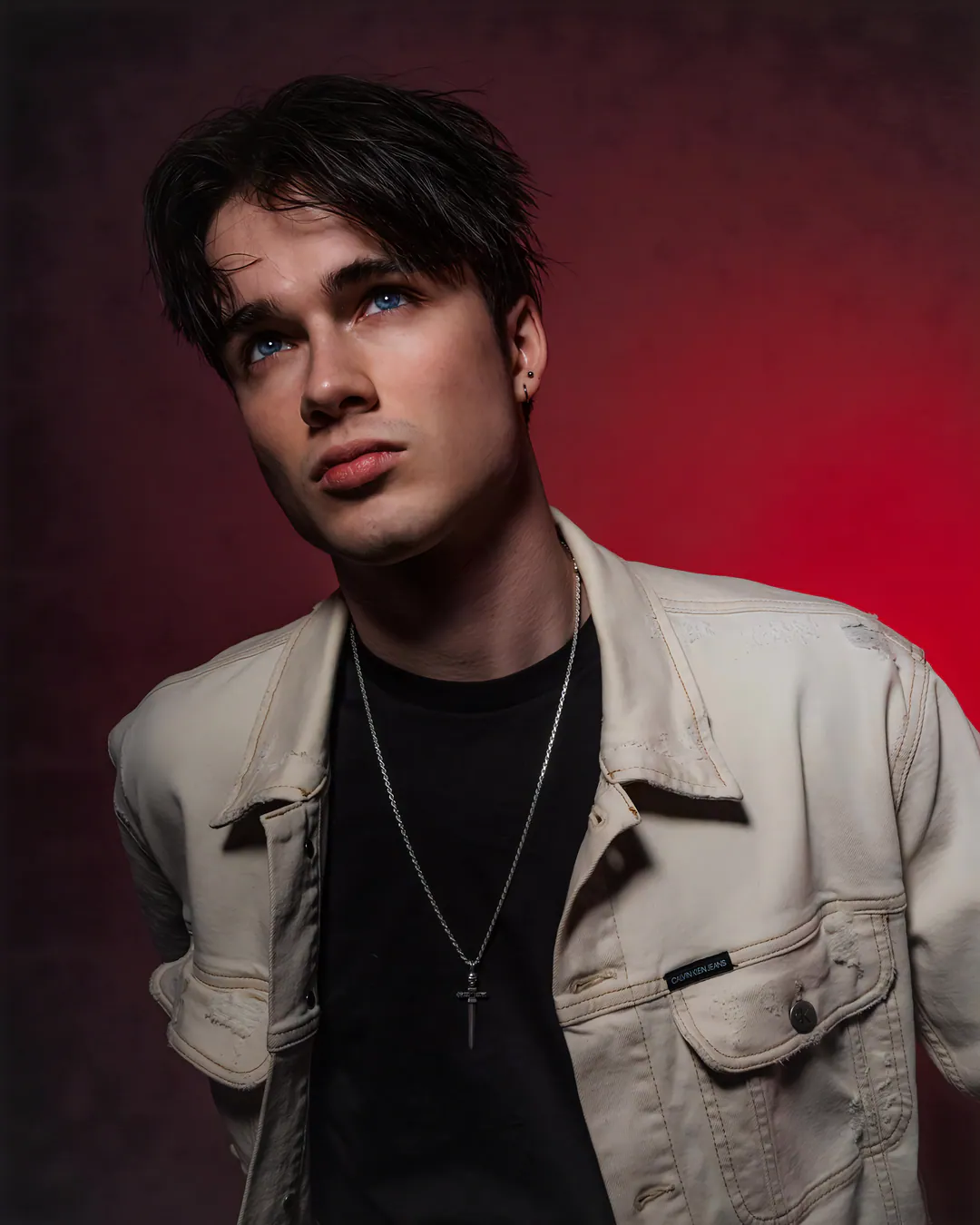 Man with tousled dark hair in a cream denim jacket and cross pendant looking up against red light