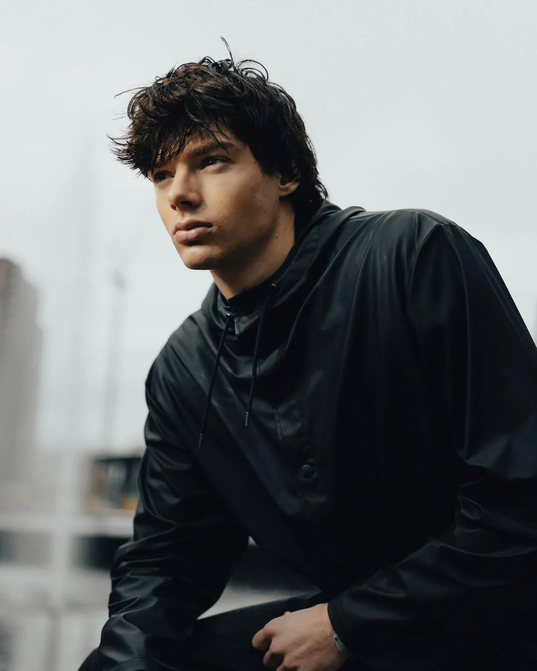 Man in a black hooded rain jacket crouching and gazing into the distance against a pale grey sky
