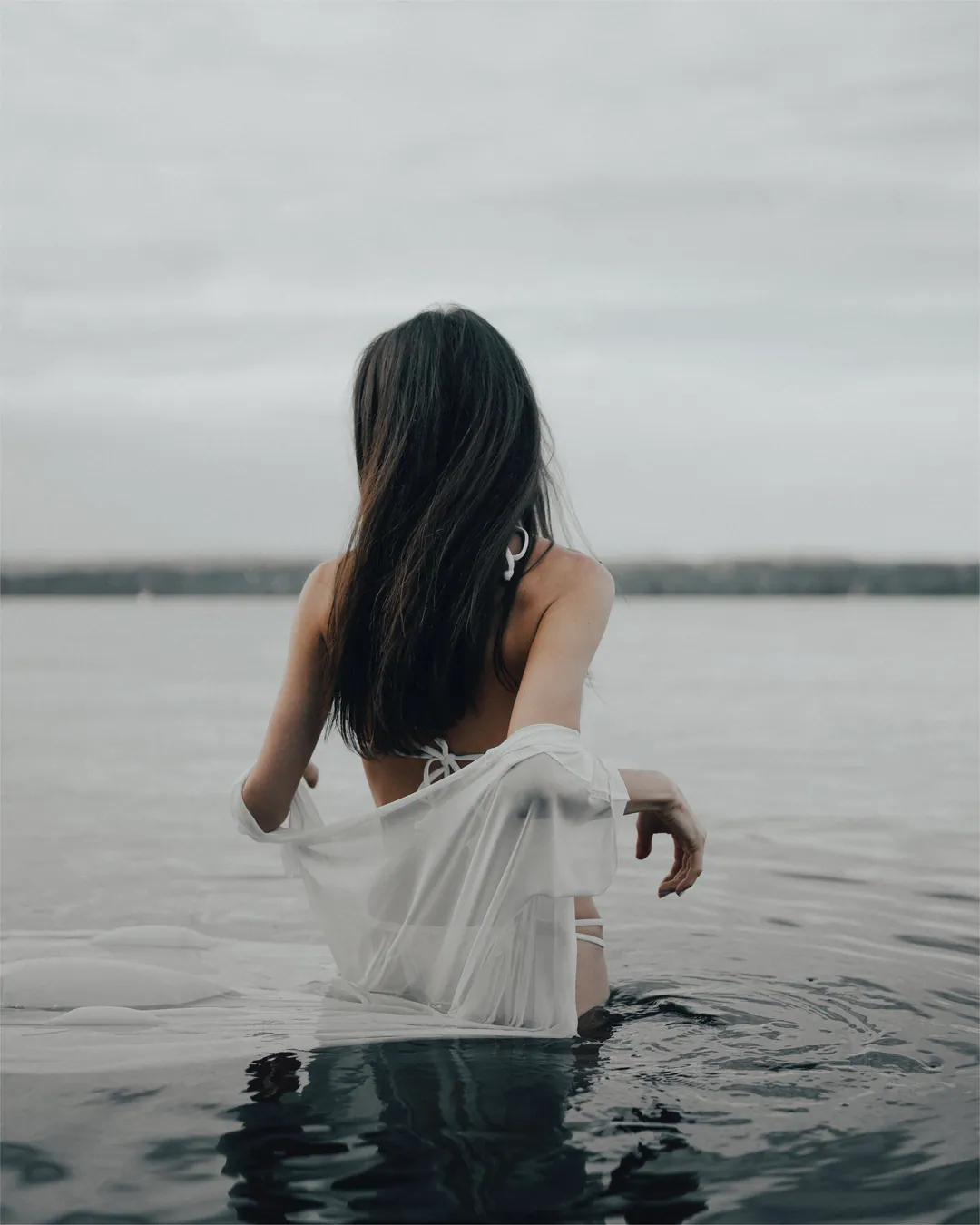 Woman with long dark hair in a sheer white wrap wading through grey water, seen from behind
