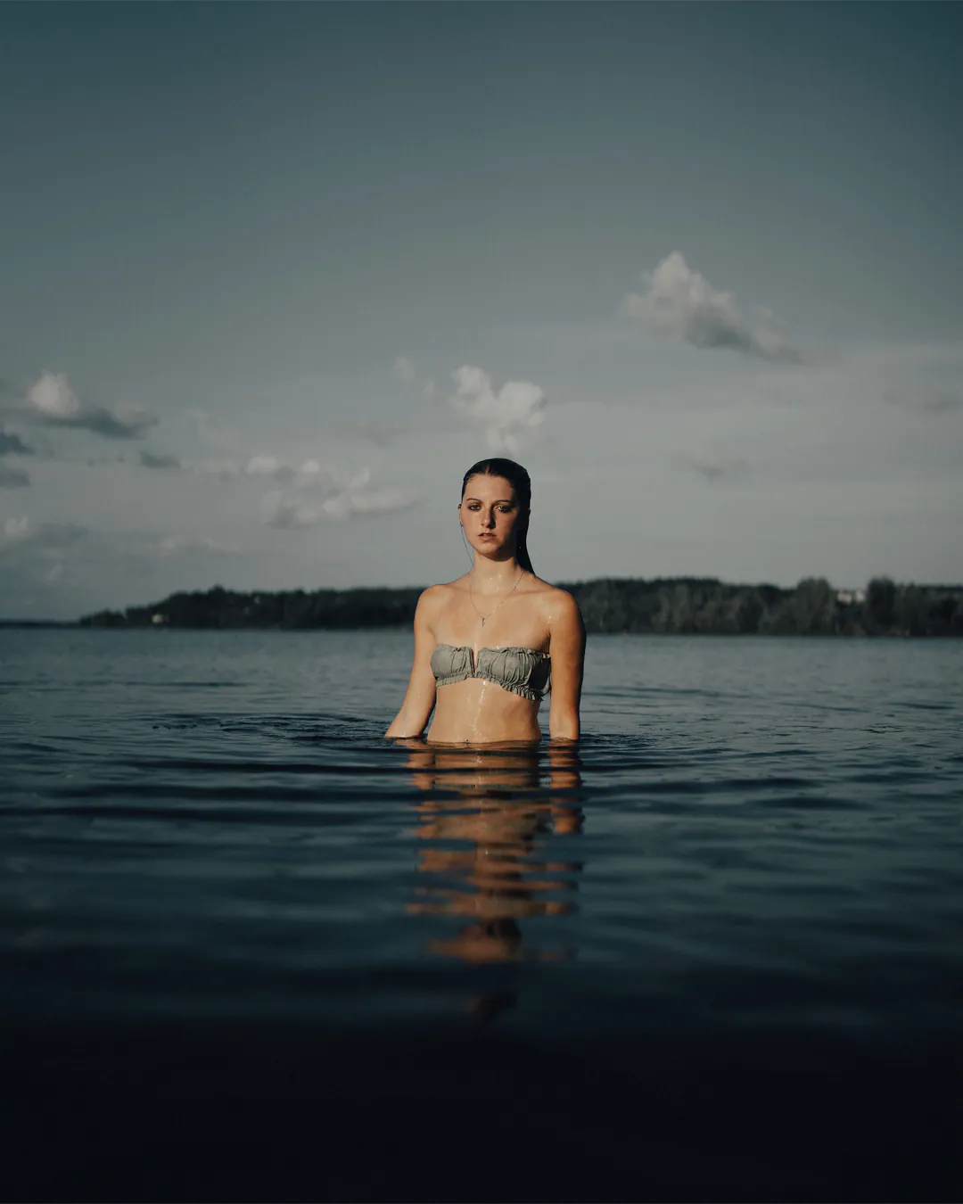 Woman in a ruched bandeau bikini top standing waist-deep in dark, still water under a dusky sky