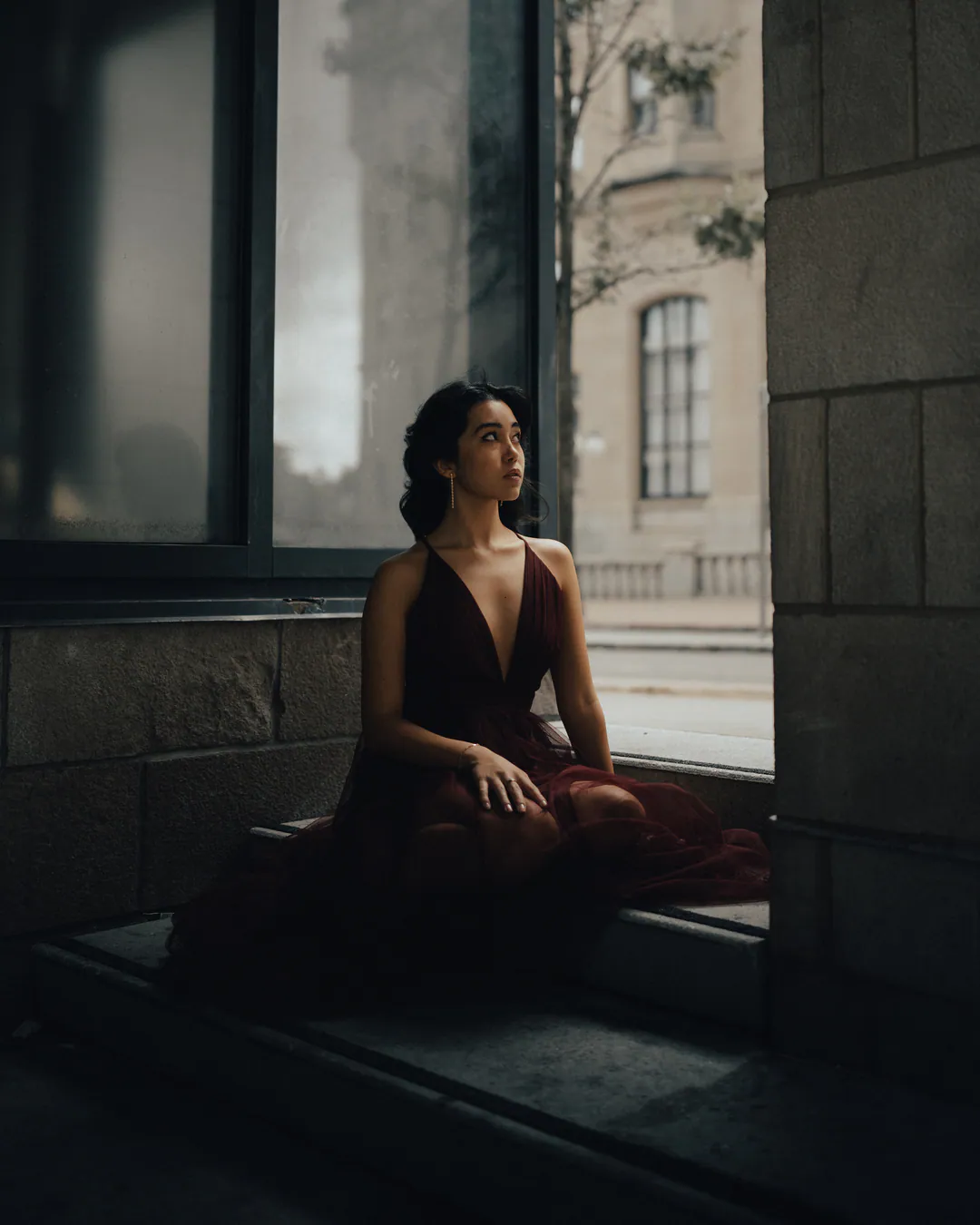 Woman in a black plunging dress sitting by a tall window in a dim stone interior
