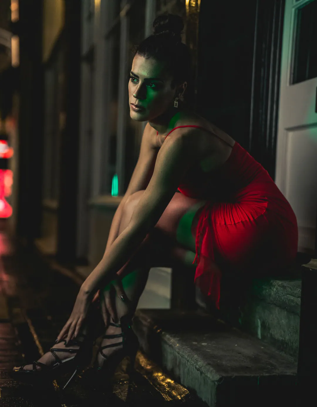 Woman in a red dress and strappy heels seated on a stone step in green and red neon