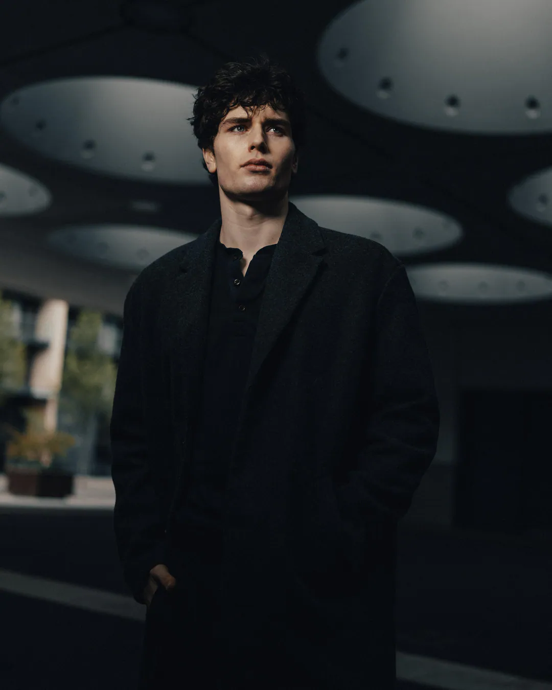 Man in a long black coat standing in a shadowy concrete undercroft beneath circular ceiling openings
