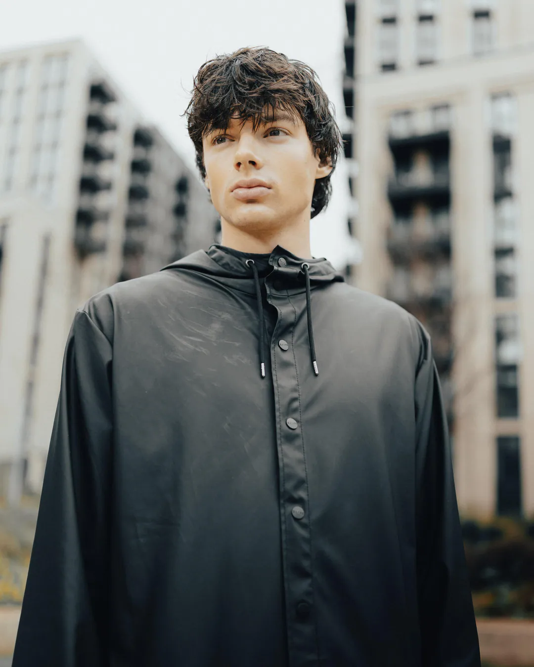 Man in a black hooded rain jacket standing between modern apartment buildings under an overcast sky