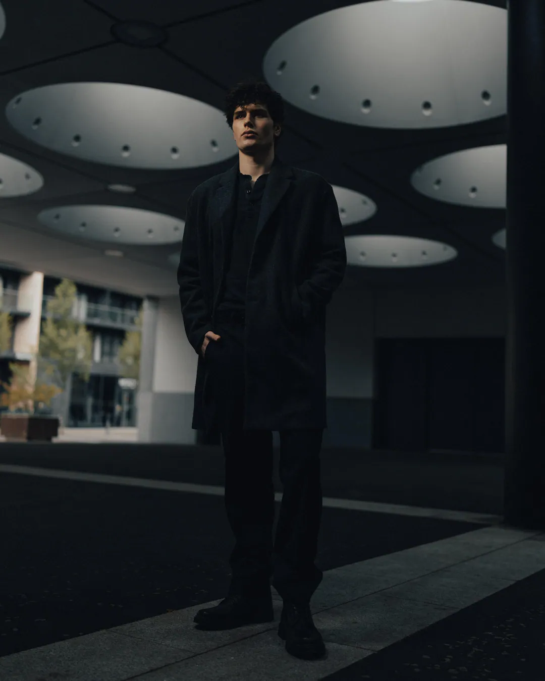 Man in a black coat standing with a hand in his pocket under a ceiling of circular skylights