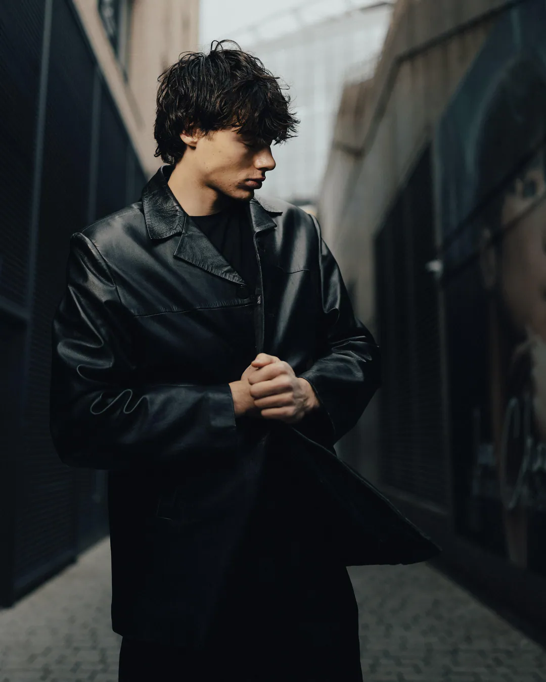 Man in a black leather coat standing in a narrow city passage, hands clasped, looking to one side