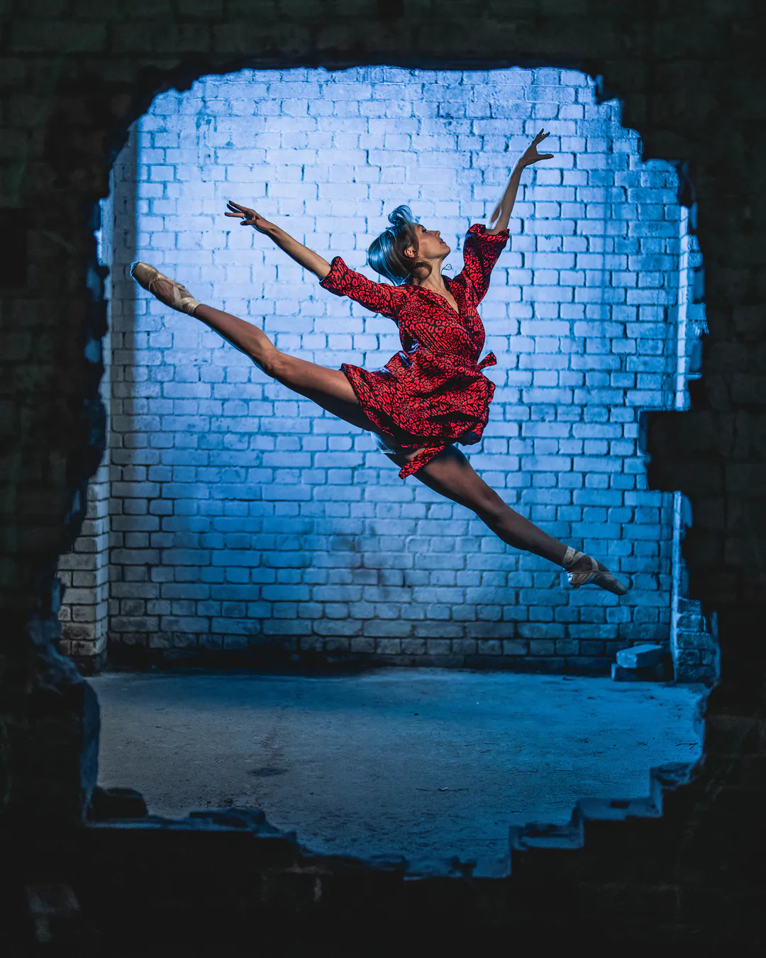 Dancer in a red ruffled dress mid-leap against a blue-lit brick wall, framed by a jagged opening
