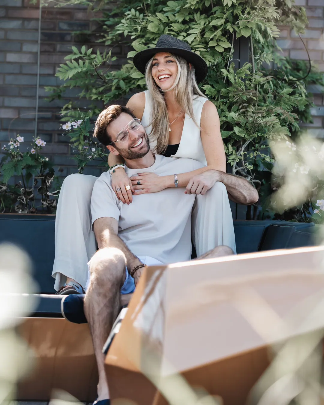 Couple laughing on a terrace bench, the woman in a black hat hugging the man from behind
