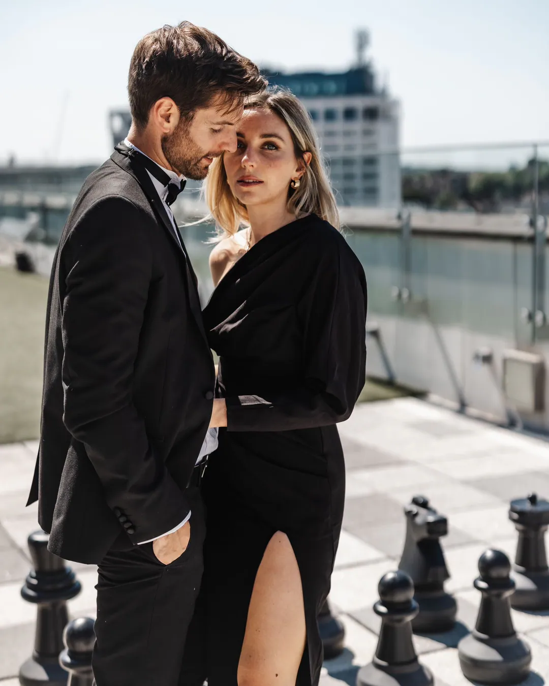 Couple in black evening wear standing close on a rooftop terrace beside giant chess pieces