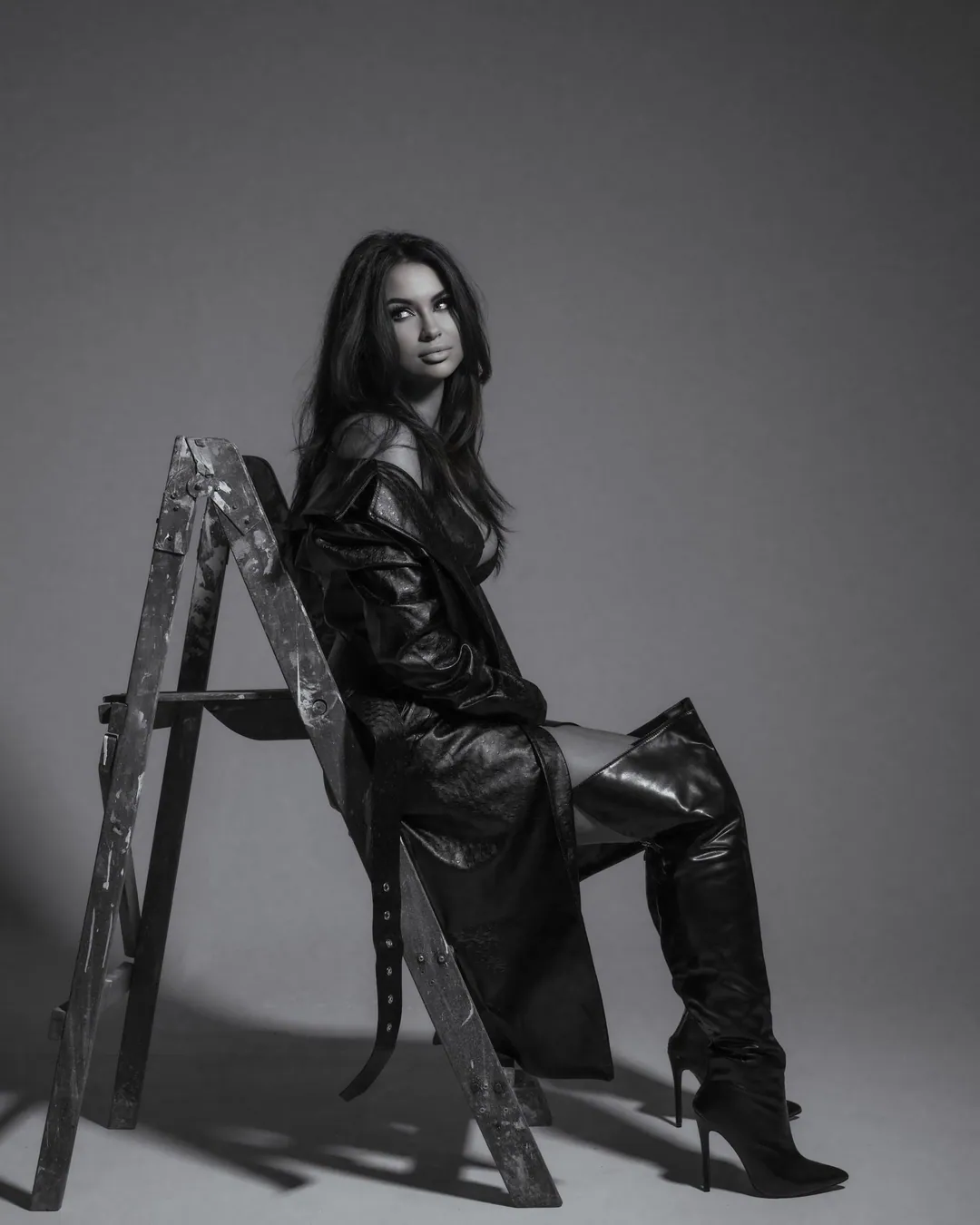 Woman in a long leather coat and thigh-high boots sitting on a paint-worn stepladder, in black and white