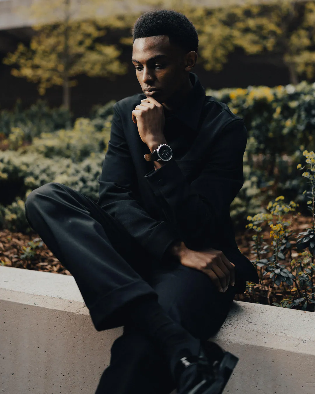 Man in black clothing sitting on a low concrete wall among shrubs, chin resting on his hand