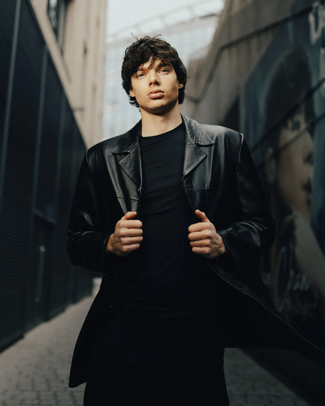 Man in a black leather coat holding its lapels in a narrow alley, shot from below