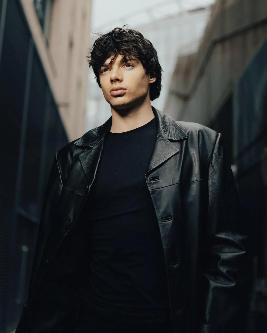 Man in a black leather coat and black T-shirt looking upwards in a narrow alley, low-angle view
