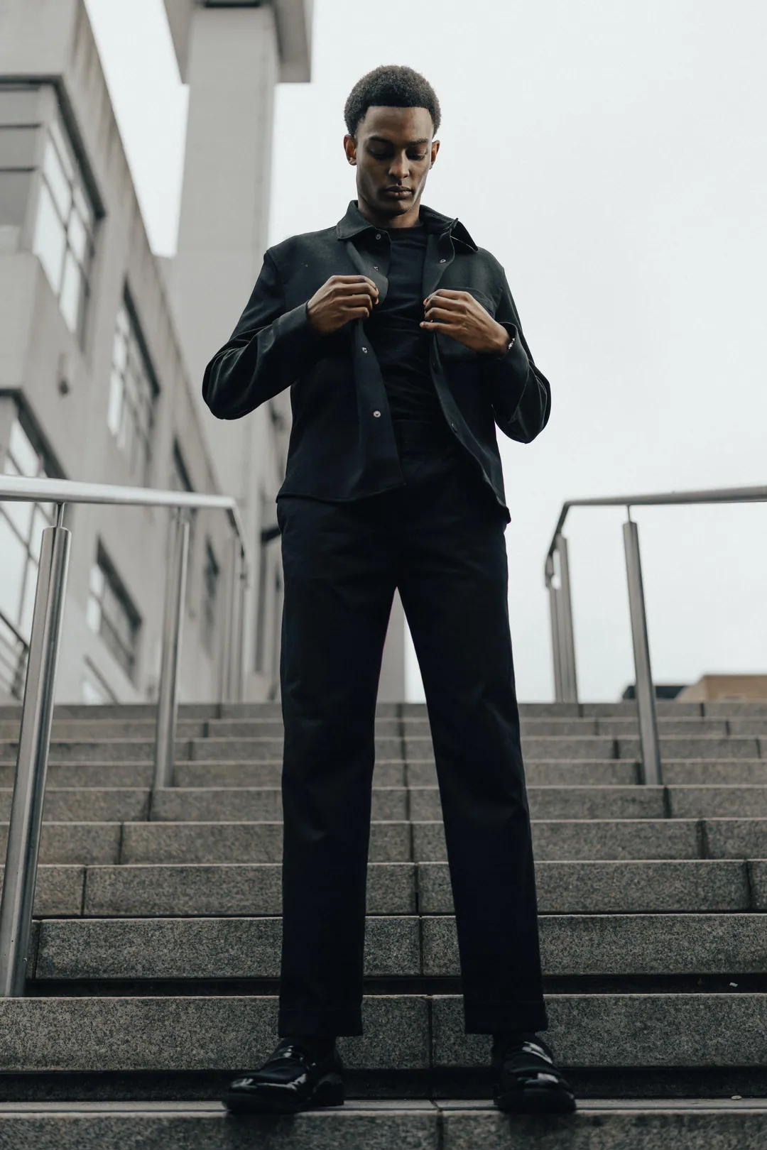 Man in a black overshirt and trousers standing on granite steps, fastening a button, shot from below