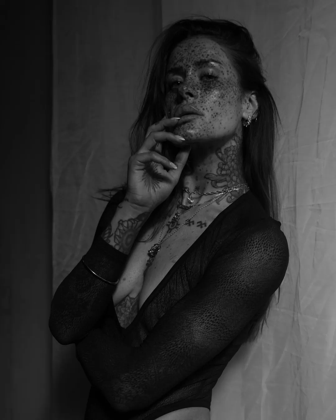 Black and white portrait of a freckled, tattooed woman in a sheer black bodysuit and fine chains, fingertips at her lip