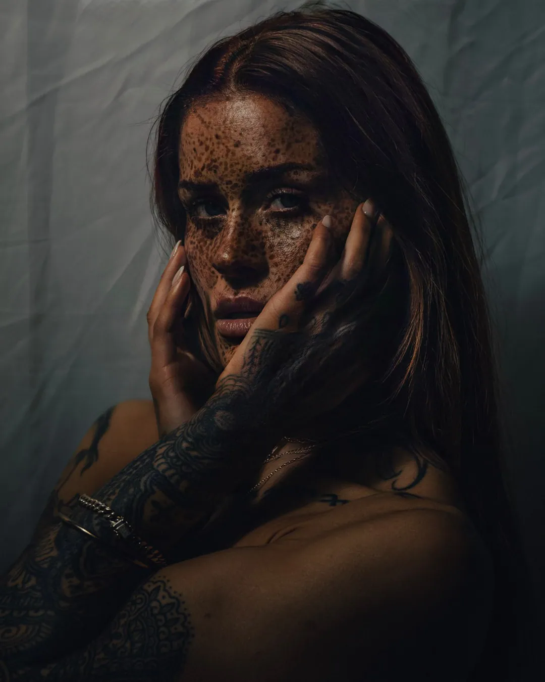 Close portrait of a heavily freckled woman cradling her face in tattooed hands against grey fabric
