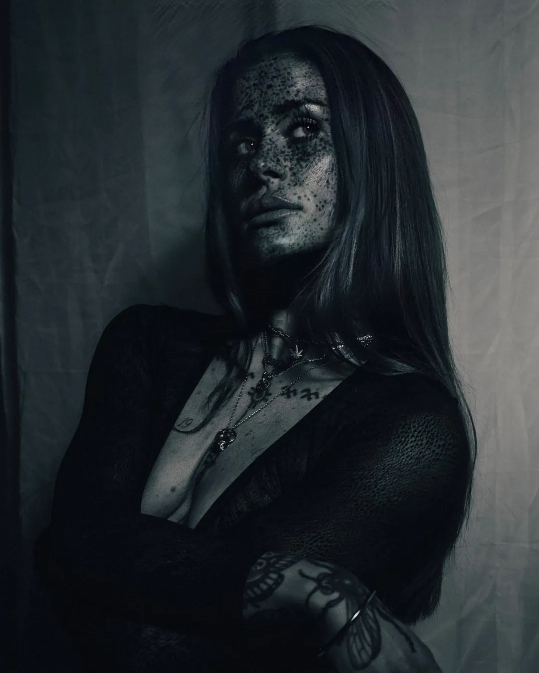 Blue-toned portrait of a freckled woman with long dark hair and layered necklaces, arms folded, glancing aside