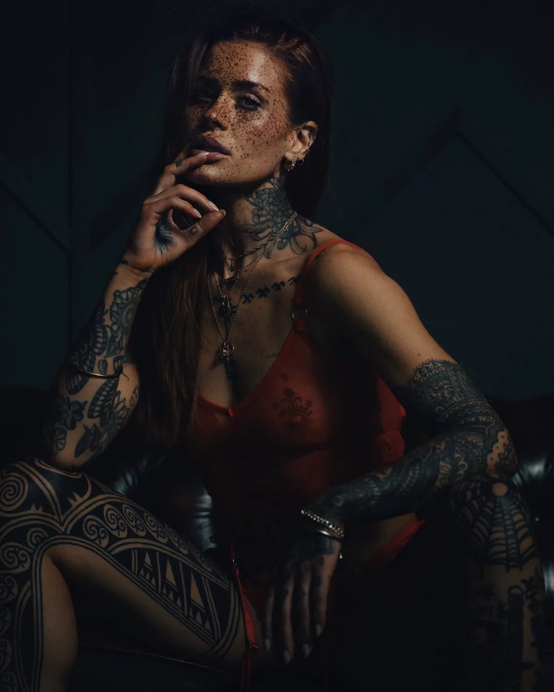 Freckled woman with tattooed arms, neck and thigh seated in a sheer red slip, fingers at her lip, in a dark room