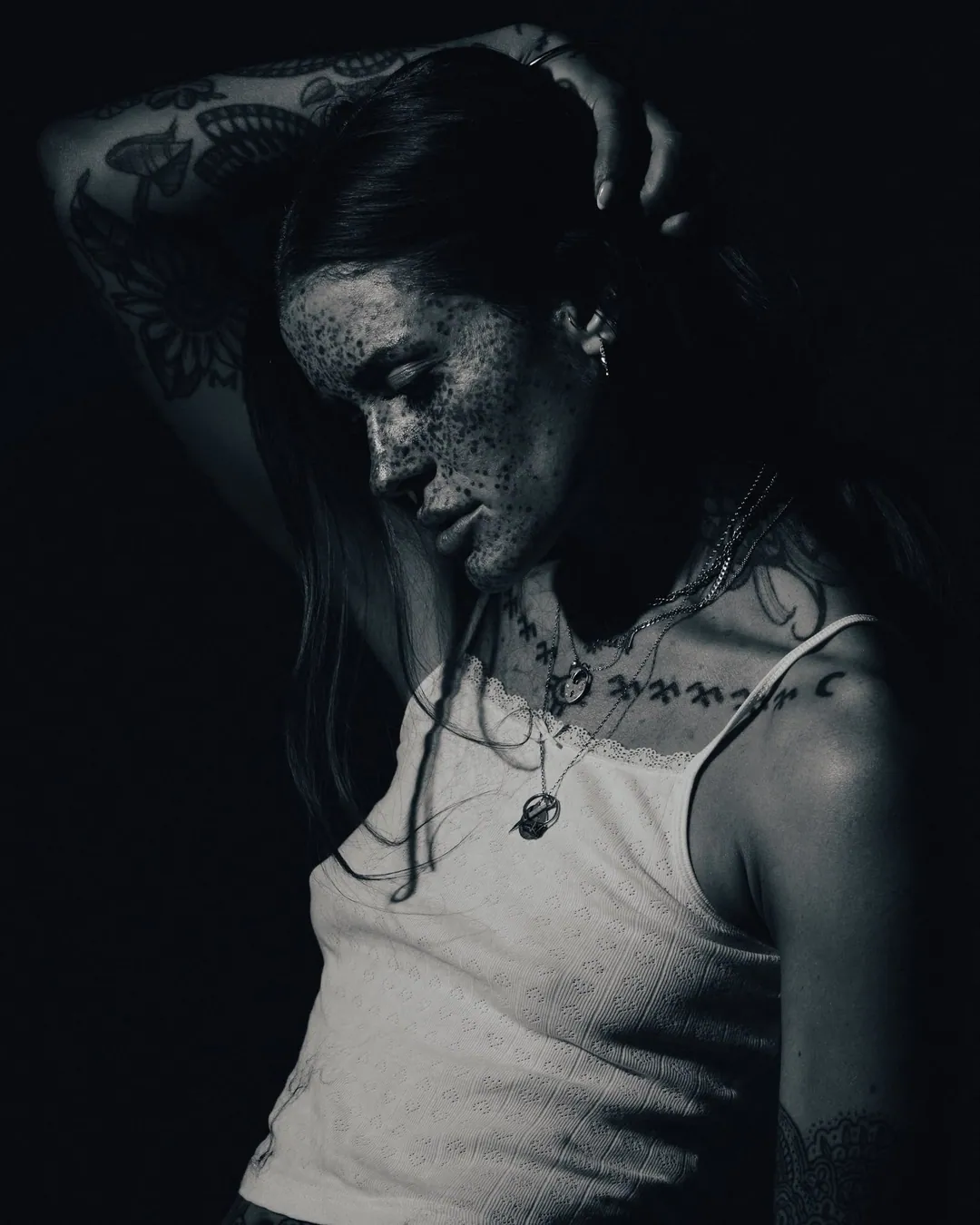 Black and white portrait of a freckled, tattooed woman in a white camisole, arm raised over her head, eyes lowered