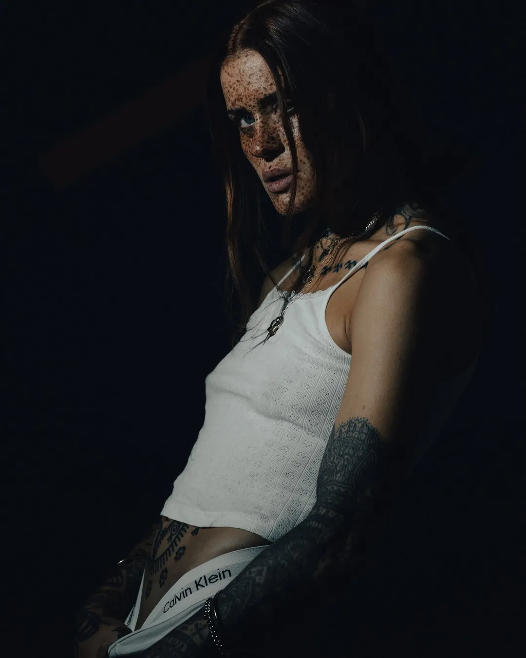 Freckled woman in a white camisole and logo waistband leaning into a pool of light, tattooed arm in shadow, against black
