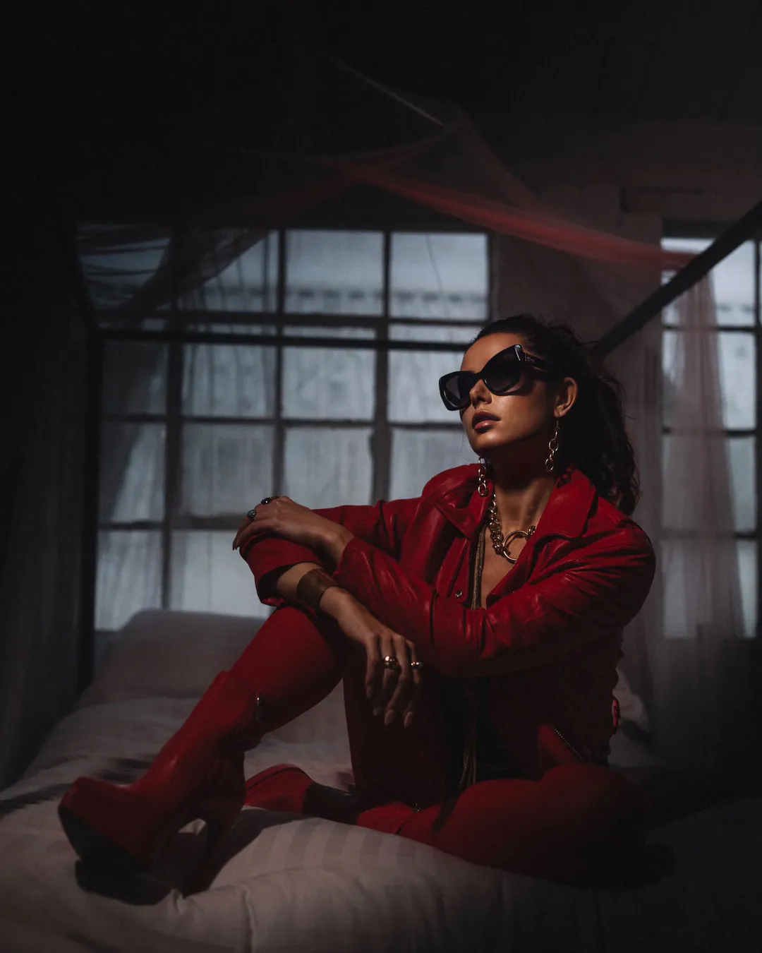 Woman in a red leather jacket, red trousers and red platform boots sitting on a canopy bed before tall loft windows, wearing black cat-eye sunglasses