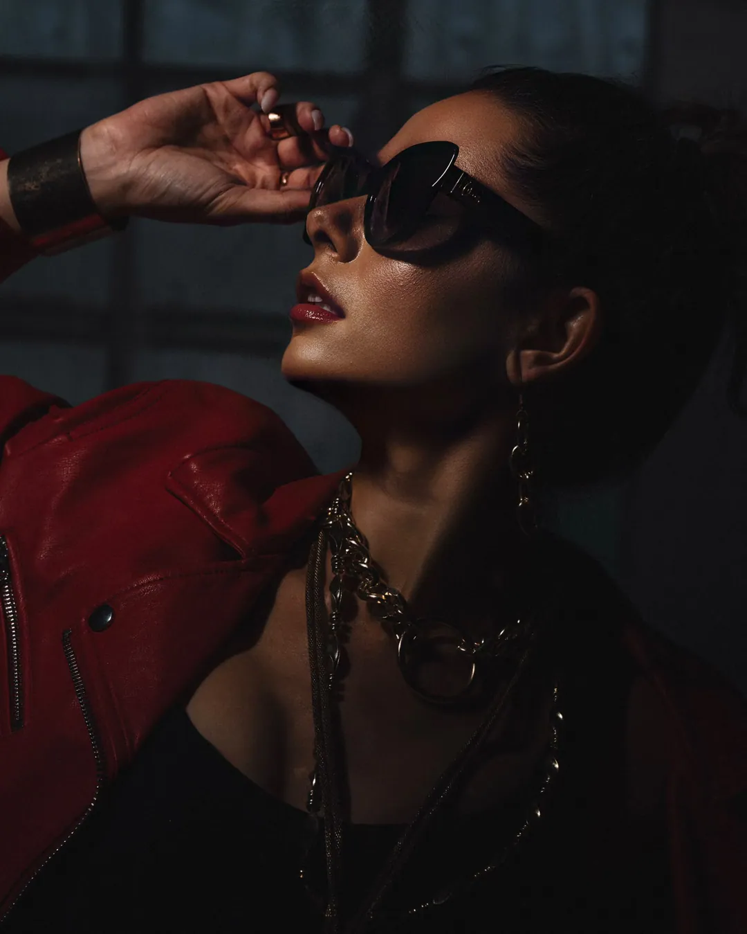 Profile of a woman in a red leather jacket and gold chains lifting the edge of her black cat-eye sunglasses, a metal cuff on her wrist