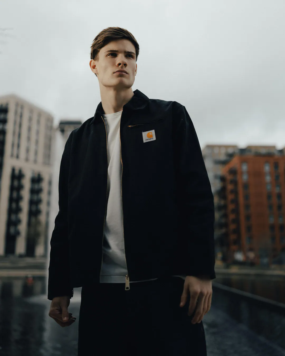 Man in a black workwear jacket standing beside a fountain among modern apartment blocks, low-angle view